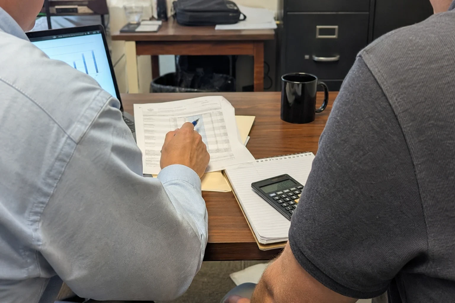 Over-the-shoulder view of two people at an office desk reviewing a printed statement with a pen, beside a notepad, calculator, black mug and filing cabinet.