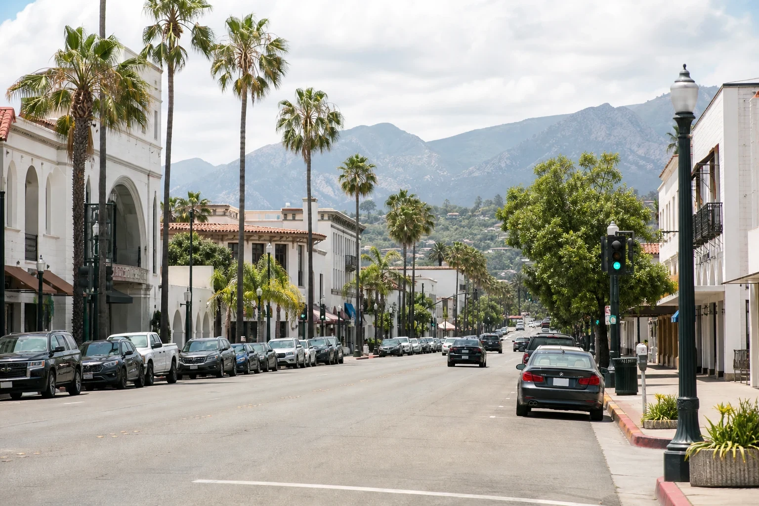 Palm-lined main street with white Spanish-style buildings, parked cars along both curbs, a green traffic light and mountains under high clouds